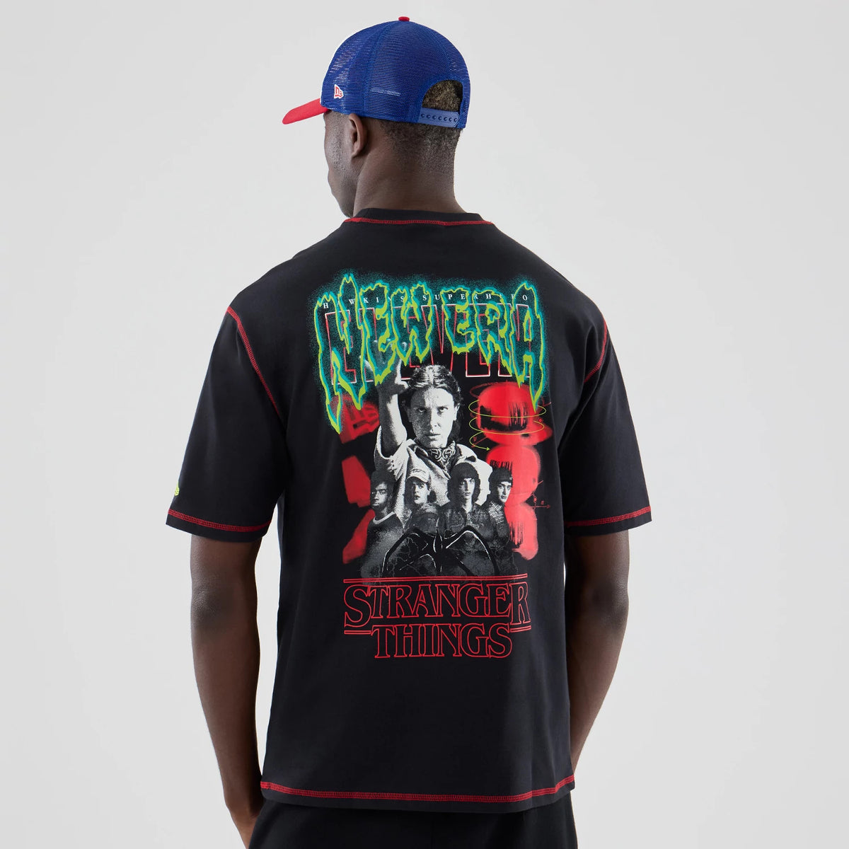 New Era Stranger Things Black Oversized T-Shirt, back view on a model with a colourful cap, featuring a detailed graphic with green New Era lettering above a collage of Stranger Things characters and red series title at the bottom.