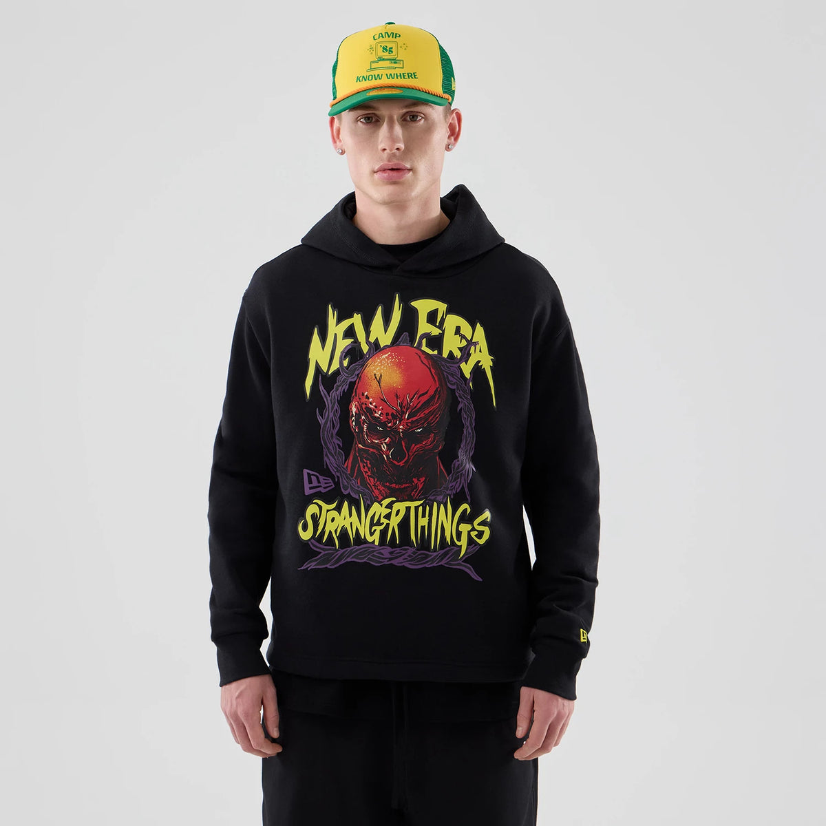 New Era Stranger Things Black Oversized Pullover Hoodie, front view on-model showing a graphic of a red demonic face below vivid yellow New Era and Stranger Things lettering, with a small embroidered logo on the left cuff.