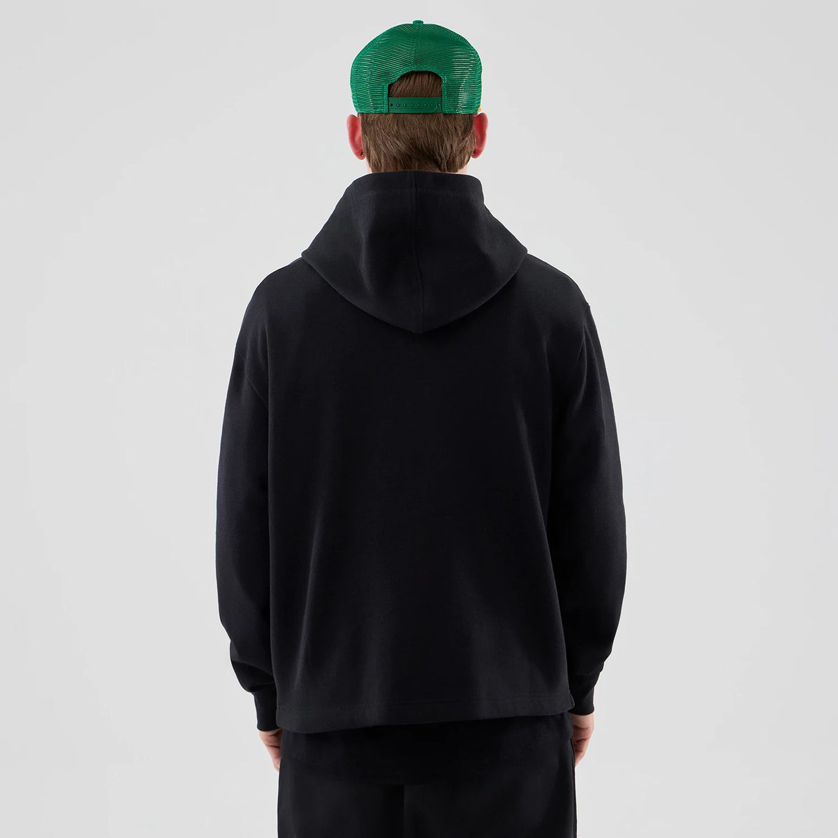 New Era Stranger Things Black Oversized Pullover Hoodie, back view on-model showing the relaxed fit of the black hoodie with a large hood and long sleeves, styled with a green cap worn backwards.