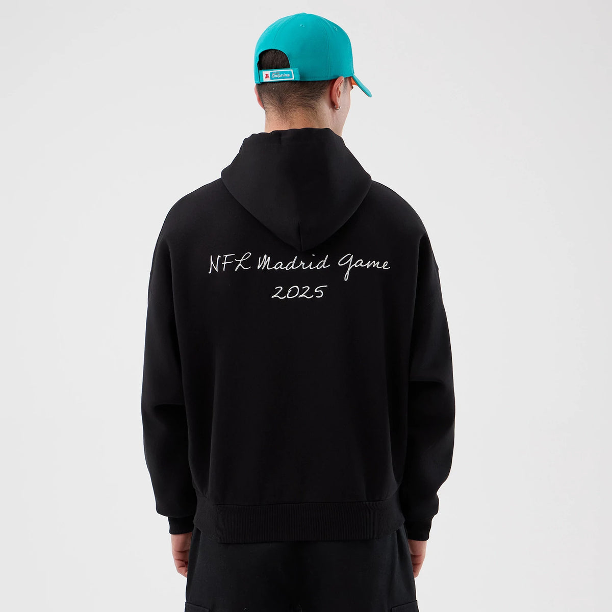 New Era Miami Dolphins NFL International Series Games Black Pullover Hoodie, back view showing NFL Madrid Game 2025 lettering across the upper back, with the hood resting flat, on-model styled with a teal cap.