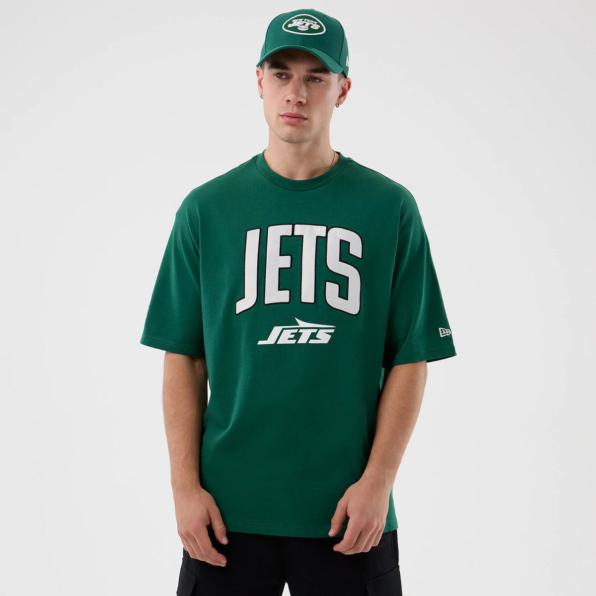 New Era New York Jets NFL International Series Games Green Oversized T-Shirt, front view on a model wearing a matching cap, showing large white printed JETS lettering centred on the chest and the New Era logo embroidered on the left sleeve.