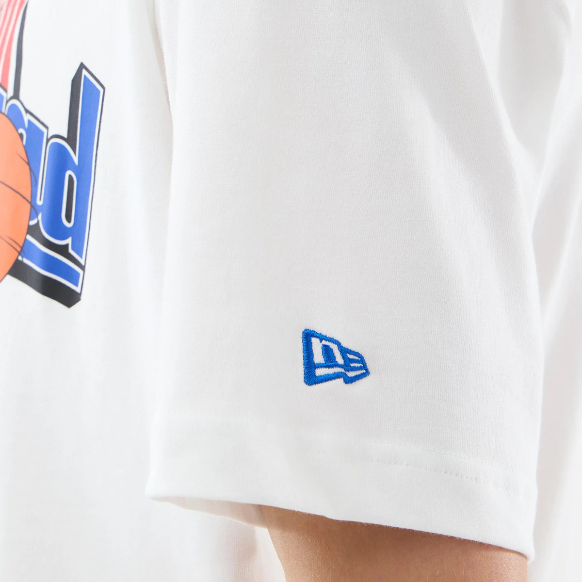 New Era New York Knicks Space Jam 2 X NBA White T-Shirt, side view showing the short sleeve with a blue embroidered New Era logo near the hem and part of a graphic with basketball elements printed on the white fabric.