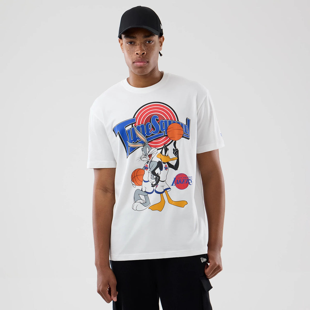 New Era LA Lakers Space Jam 2 X NBA White T-Shirt, front view on a model with a black cap, featuring a graphic of Bugs Bunny and Daffy Duck playing basketball with Tune Squad lettering and an embroidered Lakers logo near the hem.