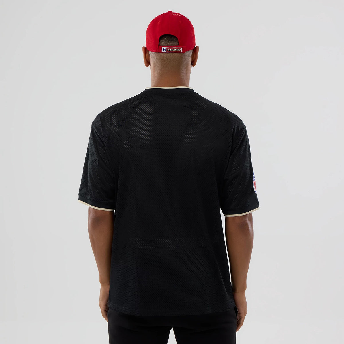 New Era San Francisco 49ers NFL Mesh Black Oversized T-Shirt, back view on-model showing the plain black mesh fabric with a loose fit, an NFL logo patch on the left sleeve and contrast beige trim around the neck and sleeve cuffs.
