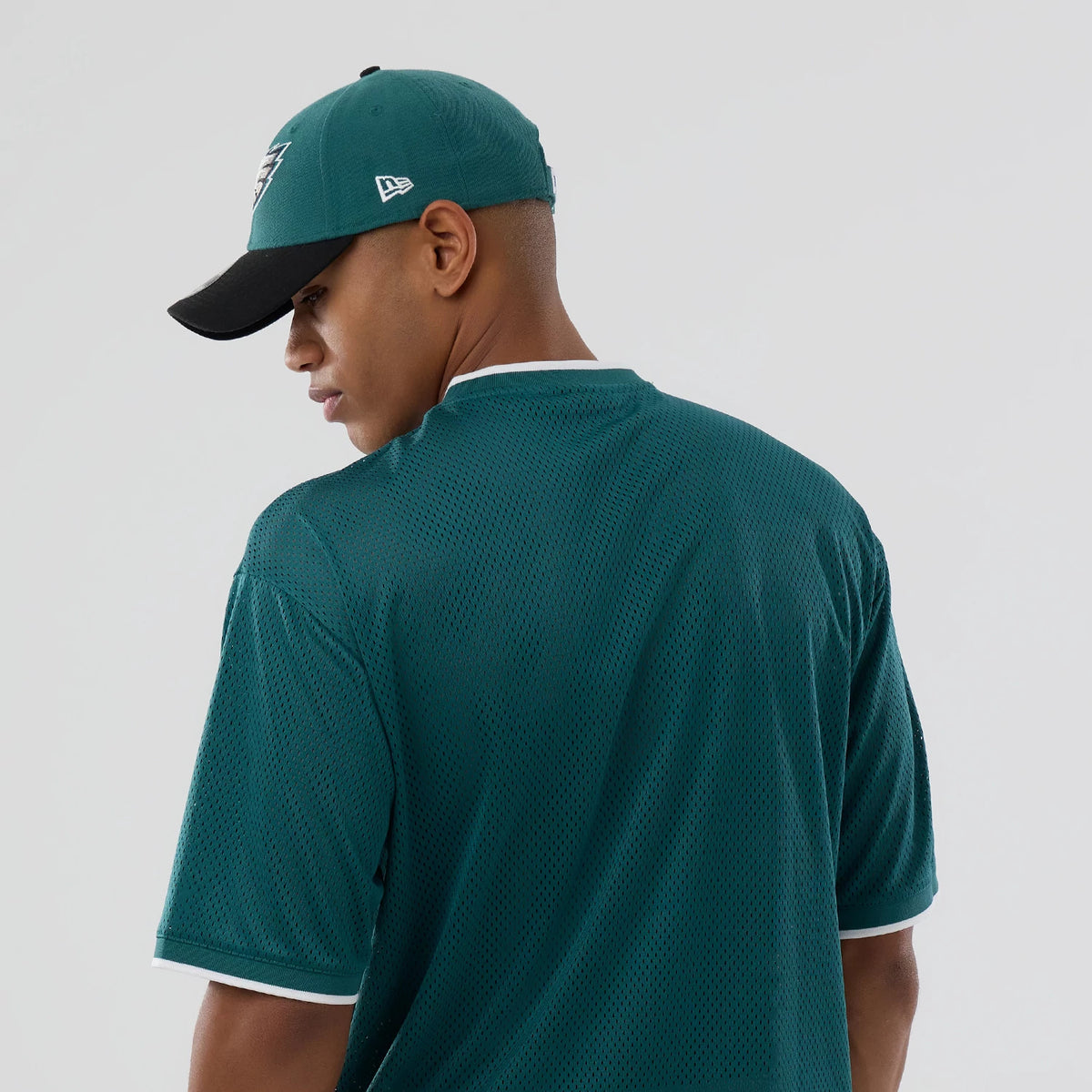 New Era Philadelphia Eagles NFL Mesh Dark Green Oversized T-Shirt, back view with a dark green mesh design and white trim on the collar and sleeves, on-model underneath a matching cap featuring an embroidered New Era logo on the side.