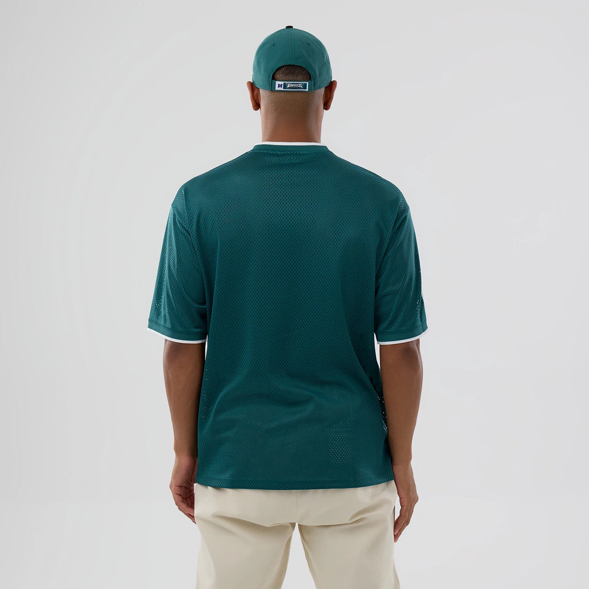 New Era Philadelphia Eagles NFL Mesh Dark Green Oversized T-Shirt, back view on a model wearing cream trousers and a matching cap, highlighting the solid dark green mesh fabric with white trims on the sleeves and collar.