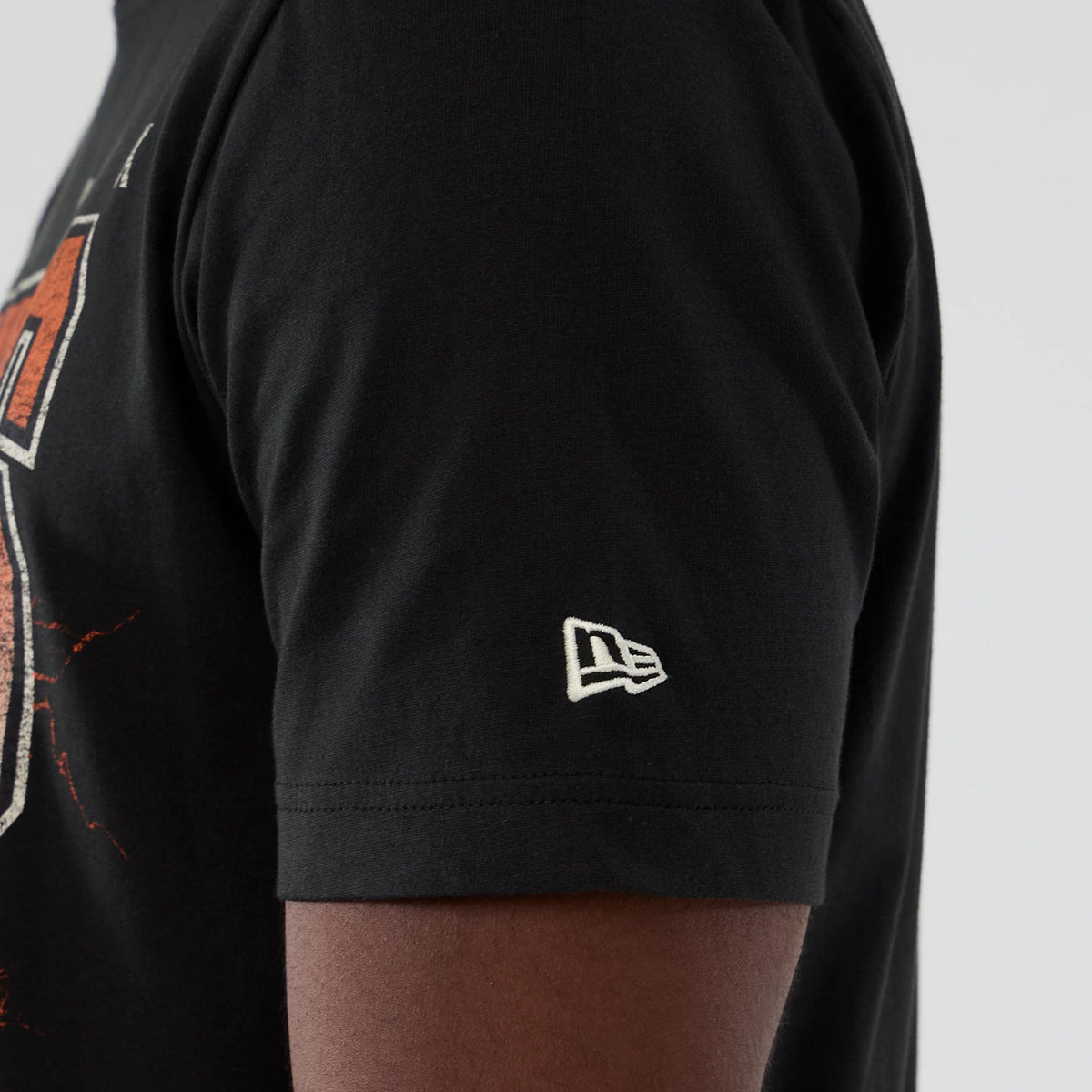 New Era New York Knicks NBA Hardwood Classic Black T-Shirt, side view showing the embroidered white New Era logo on the sleeve edge, with part of an orange and white graphic partially on the front panel.