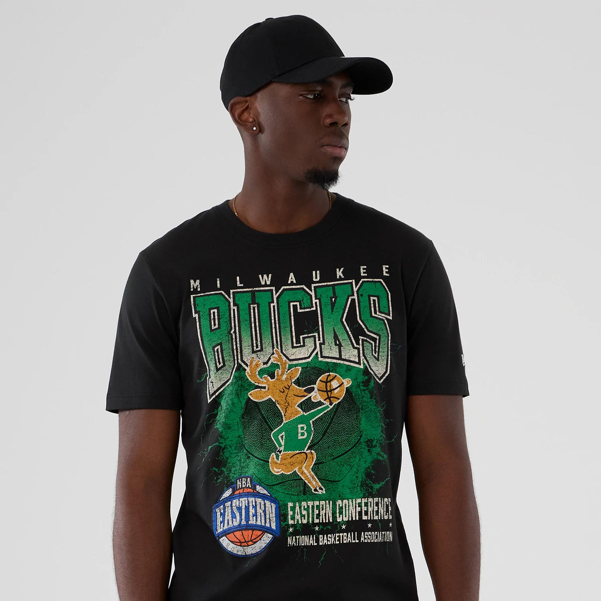 New Era Milwaukee Bucks NBA Hardwood Classic Black T-Shirt, front view on a model styled with a black cap, featuring large green and white Bucks logo, team graphics including a deer dribbling a basketball.