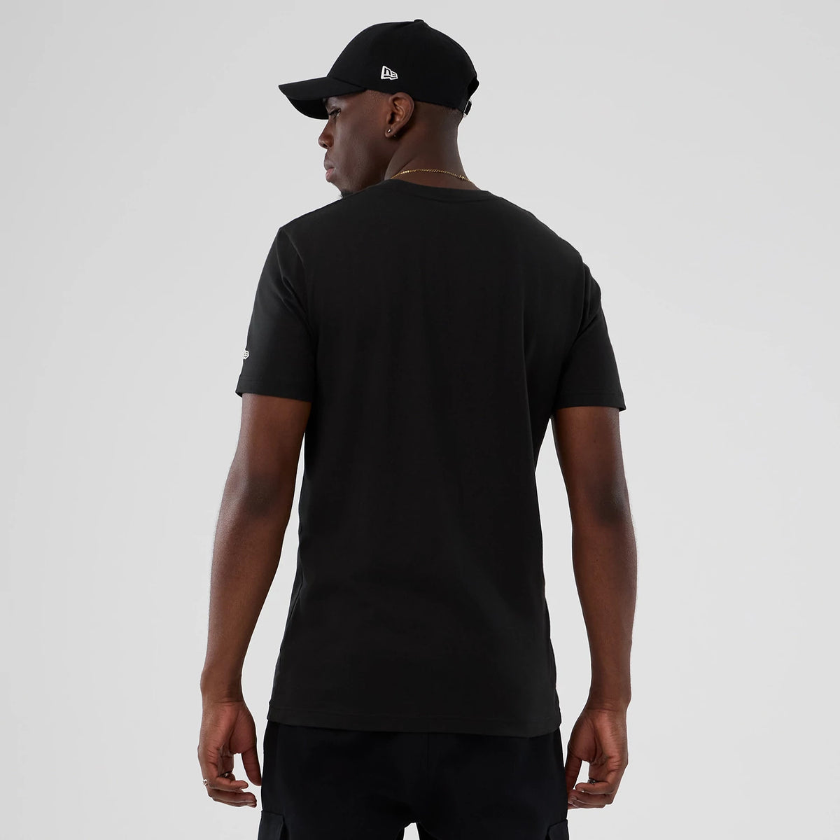 New Era Detroit Pistons NBA Hardwood Classic Black T-Shirt, back view on-model highlighting the plain black design with short sleeves, and a small embroidered New Era logo on the left sleeve paired with a black cap featuring a raised New Era logo.