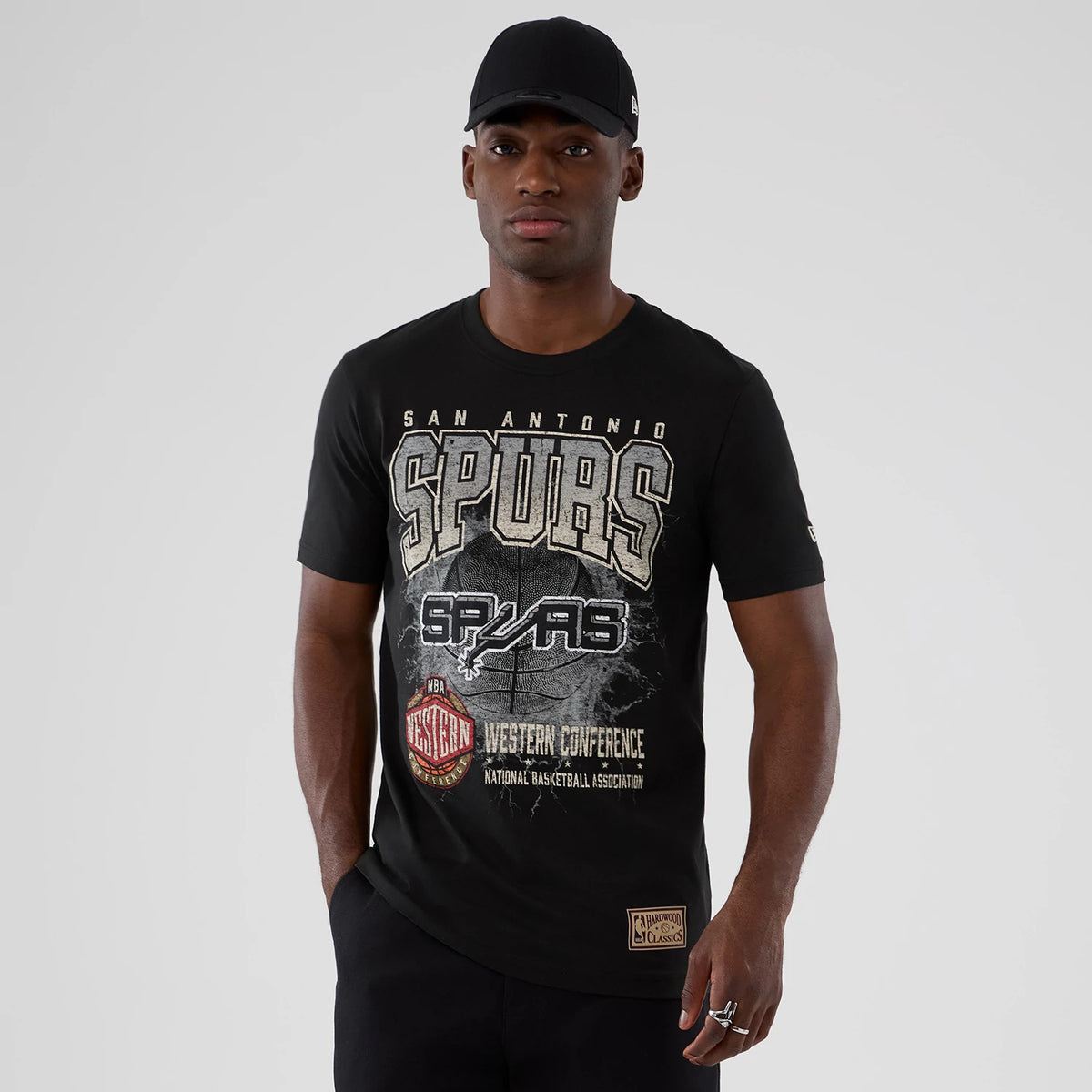 New Era San Antonio Spurs NBA Hardwood Classic Black T-Shirt, front view on a model styled with a black cap, featuring large Spurs lettering across the chest, with additional graphic details and a printed patch near the hem.