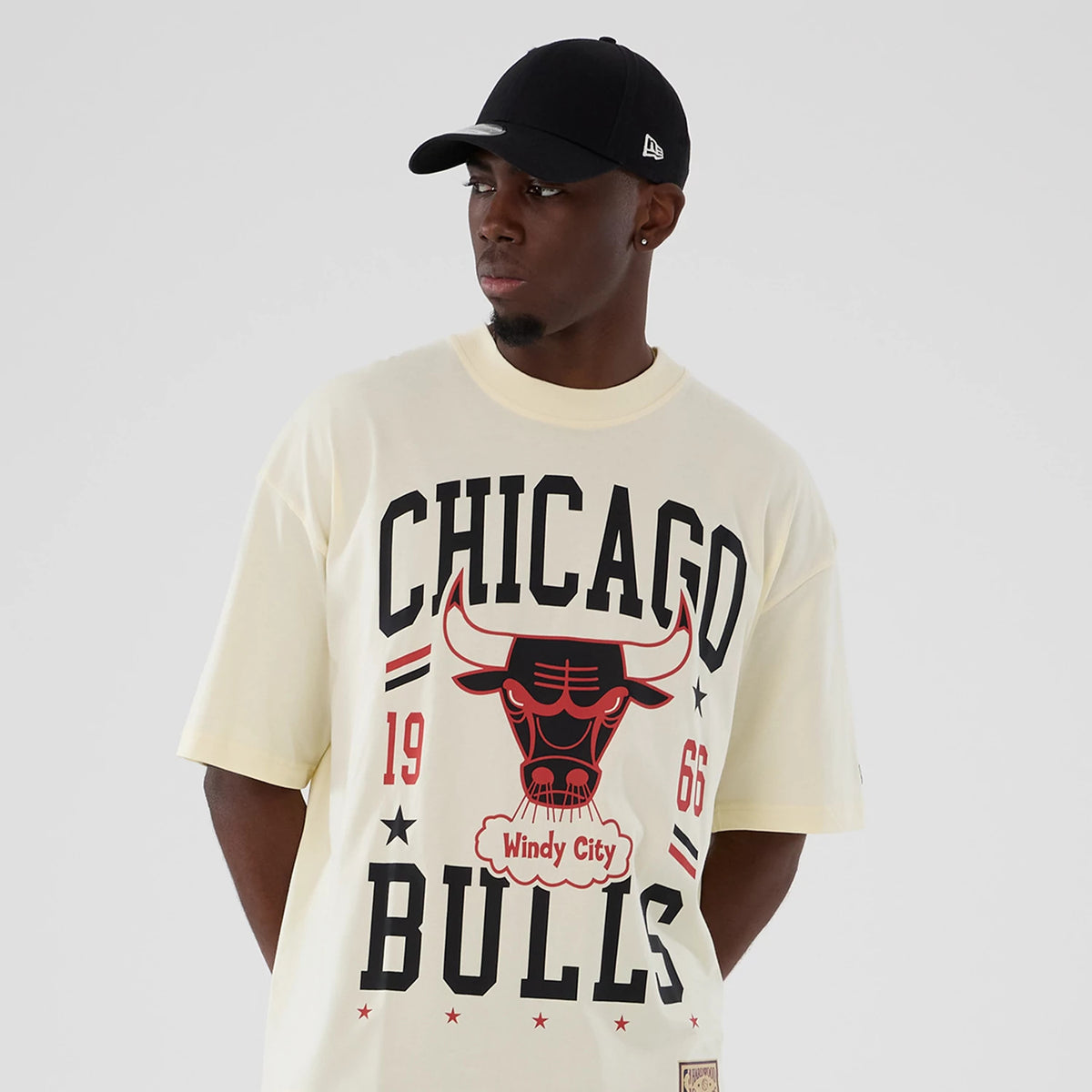 New Era Chicago Bulls NBA Hardwood Classic Cream T-Shirt, front view featuring a large printed Chicago Bulls logo with Windy City and 1966 details in black and red on a cream background, worn with a simple black cap for a casual look.