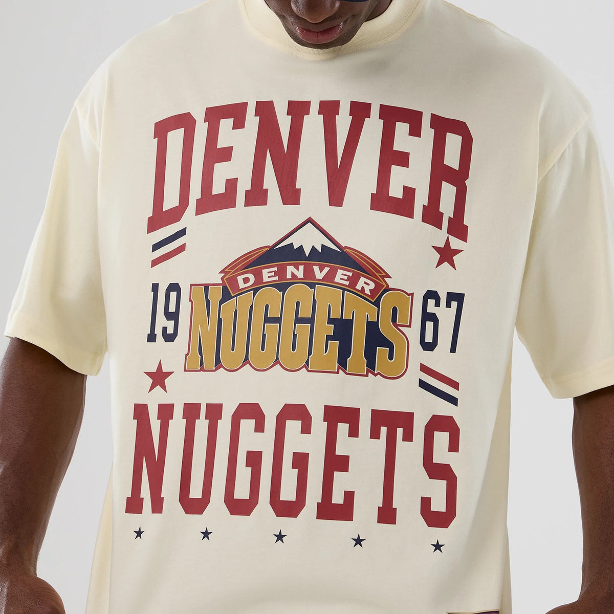 New Era Denver Nuggets NBA Hardwood Classic Cream T-Shirt, front view displaying a graphic with Denver Nuggets lettering in red and gold, a stylised mountain logo in the centre, the year 1967, and decorative stars around the design.