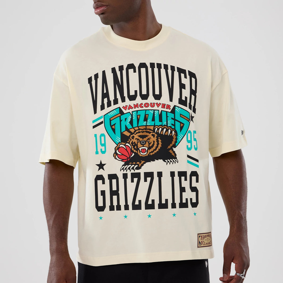 New Era Vancouver Grizzlies NBA Hardwood Classic Cream T-Shirt, front view on a model showcasing Vancouver Grizzlies graphics with a bear holding a basketball, black lettering, turquoise accents, and an embroidered New Era logo on the left sleeve.