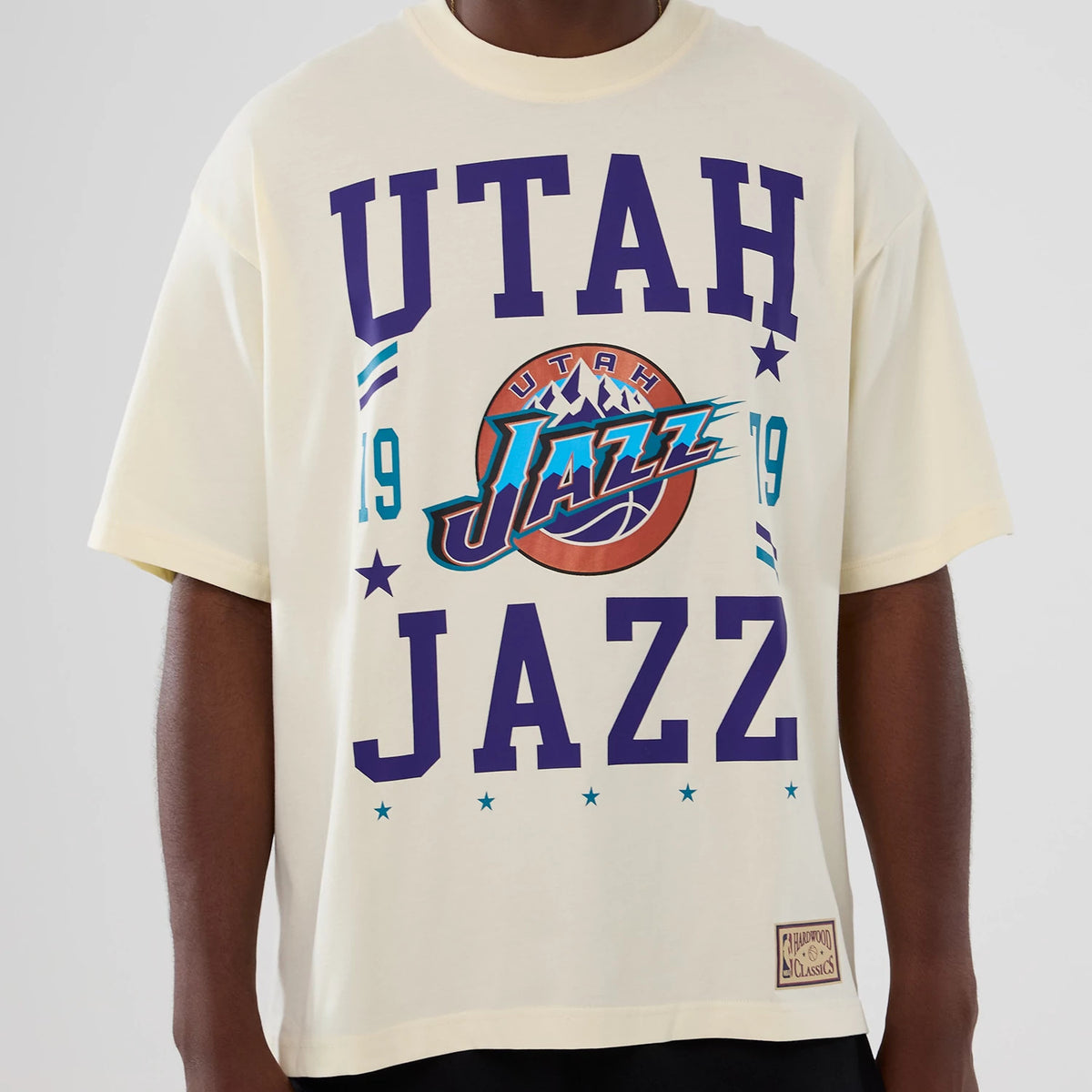New Era Utah Jazz NBA Hardwood Classic Cream T-Shirt, front view displaying purple UTAH JAZZ text and a colourful team graphic with the number 19 on the cream fabric, worn by a model with a small Hardwood Classics patch near the hem.