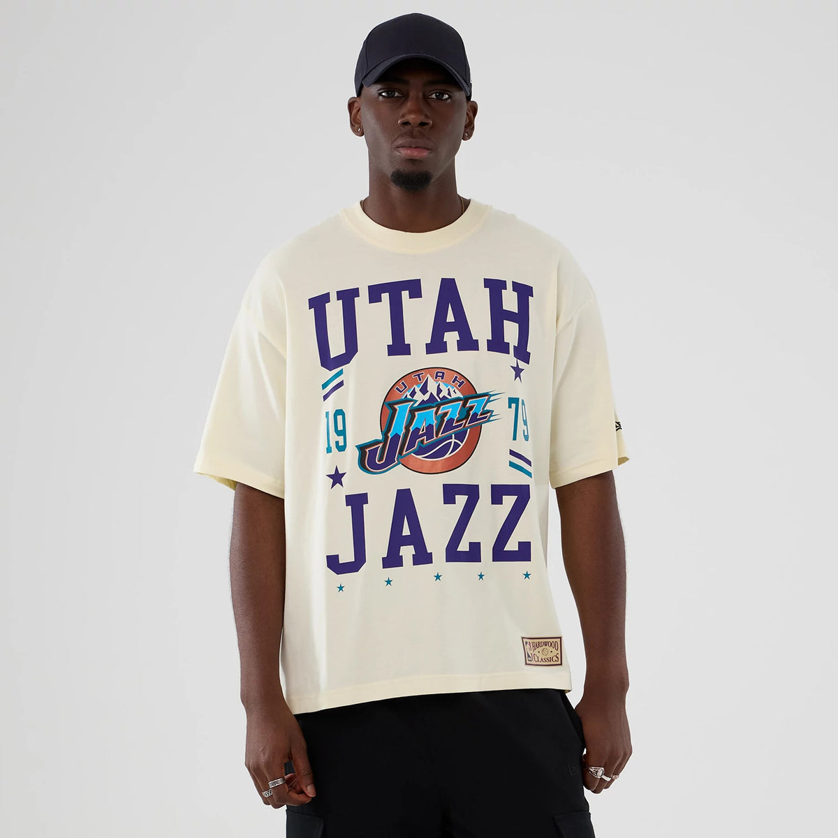 New Era Utah Jazz NBA Hardwood Classic Cream T-Shirt, front view on a model styled with a plain cap, featuring purple UT Jazz lettering and a large logo with a basketball motif centred on the chest.