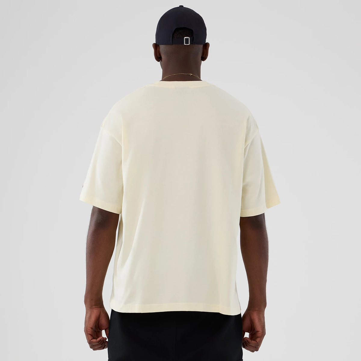 New Era Utah Jazz NBA Hardwood Classic Cream T-Shirt, back view displaying the plain cream back with short sleeves, worn casually on a model without graphics or branding, paired with dark trousers and a navy cap.