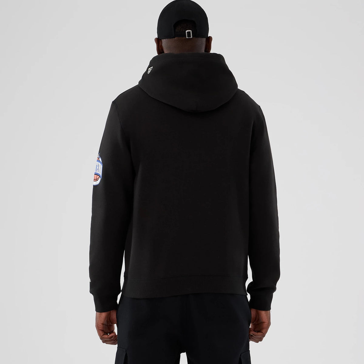 New Era Boston Celtics NBA Hardwood Classic Black Pullover Hoodie, back view on a model with a black cap, showing a small embroidered New Era logo on the hood and a detailed patch on the sleeve.