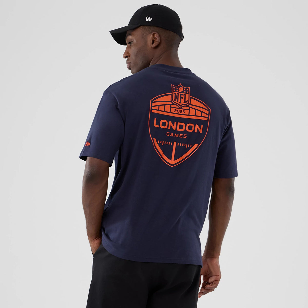 New Era Denver Broncos NFL International Series Games 2025 Navy Oversized T-Shirt, back view showing a large orange graphic with NFL 2025 London Games text, worn by a model with contrasting black shorts and a black cap.