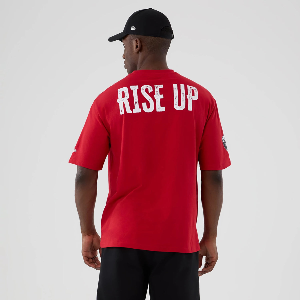 New Era Atlanta Falcons NFL International Series Games 2025 Red Oversized T-Shirt, back view on-model showing the white RISE UP lettering across the upper back, the team logo embroidered on the left sleeve and a New Era logo on the right sleeve.