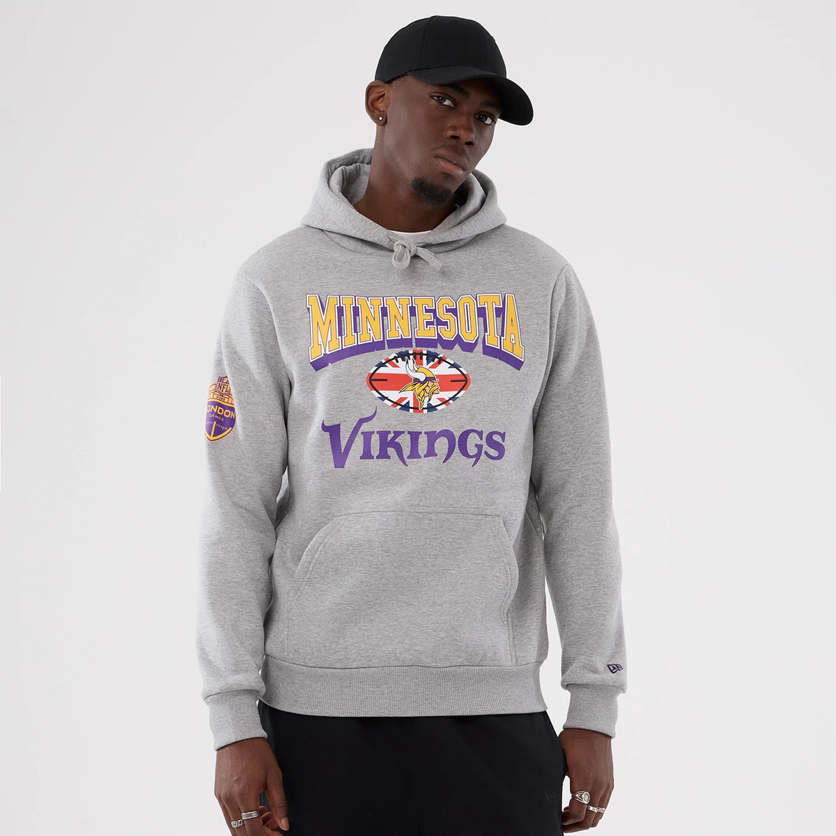New Era Minnesota Vikings NFL International Series Games 2025 Grey Pullover Hoodie, front view on a model showing the yellow Minnesota and purple Vikings lettering with a central logo, small patch on the left sleeve.
