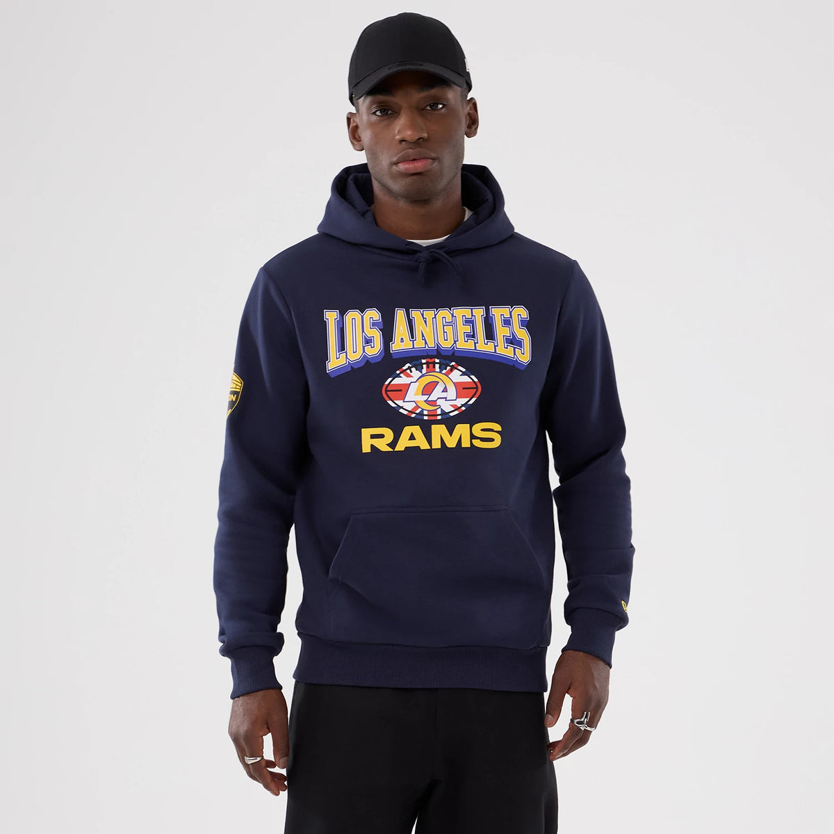 New Era LA Rams NFL International Series Games 2025 Navy Pullover Hoodie, front view on-model showing the large yellow and blue Los Angeles Rams lettering across the chest, a graphical emblem below, and a logo patch on the left sleeve.