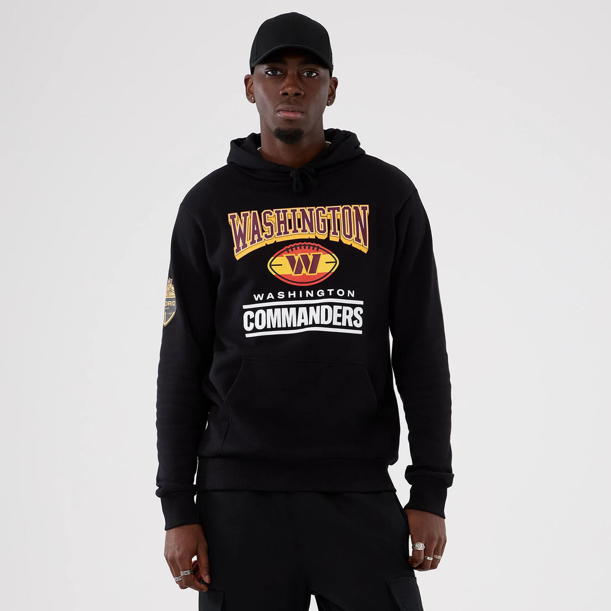 New Era Washington Commanders NFL International Series Games 2025 Black Pullover Hoodie, front view on-model with large Commanders lettering and logo across the chest, a sleeve patch on the left arm, and a drawstring hood positioned.