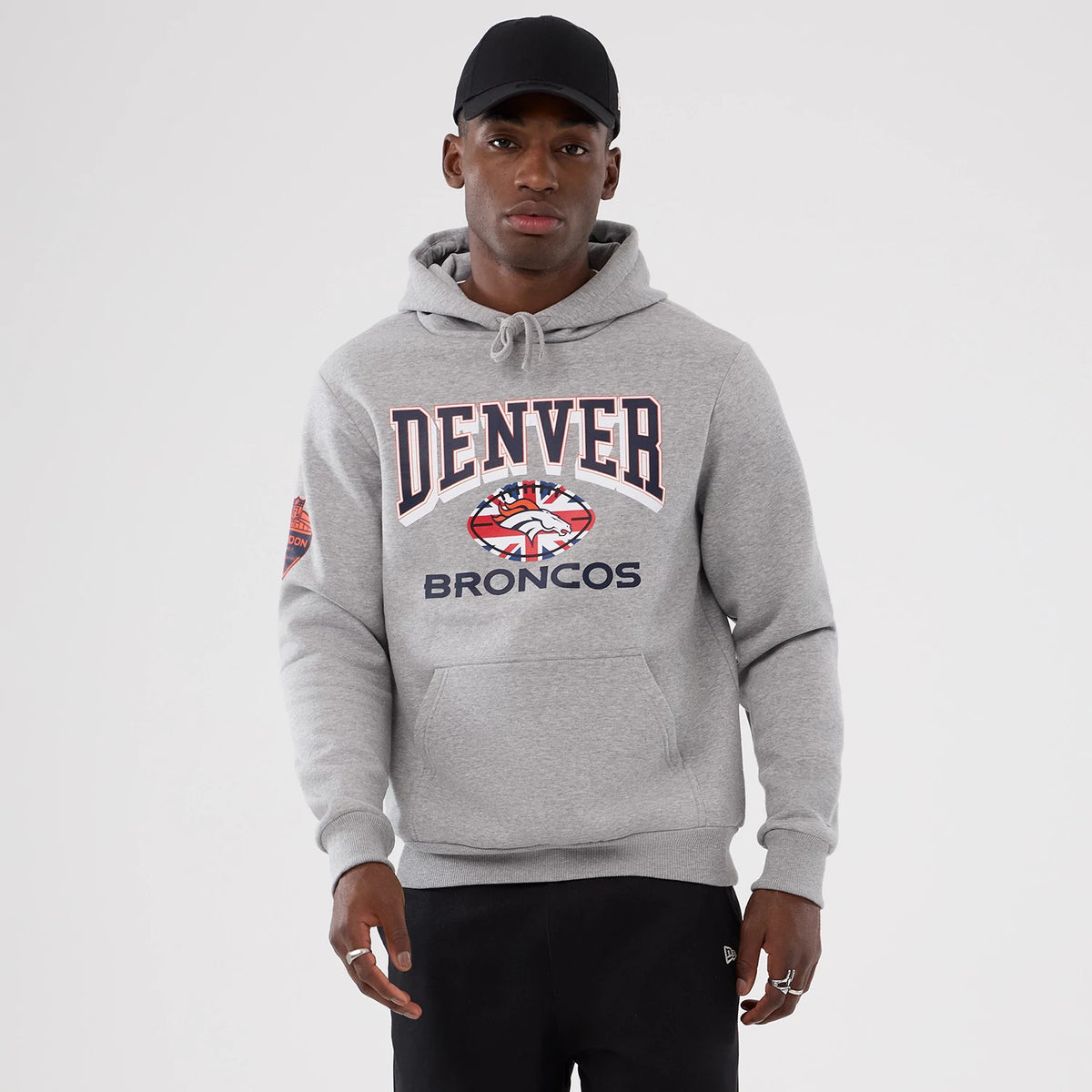 New Era Denver Broncos NFL International Series Games 2025 Grey Pullover Hoodie, front view showing the DENVER lettering across the chest with a printed Broncos logo underneath, worn by a model in a black cap.