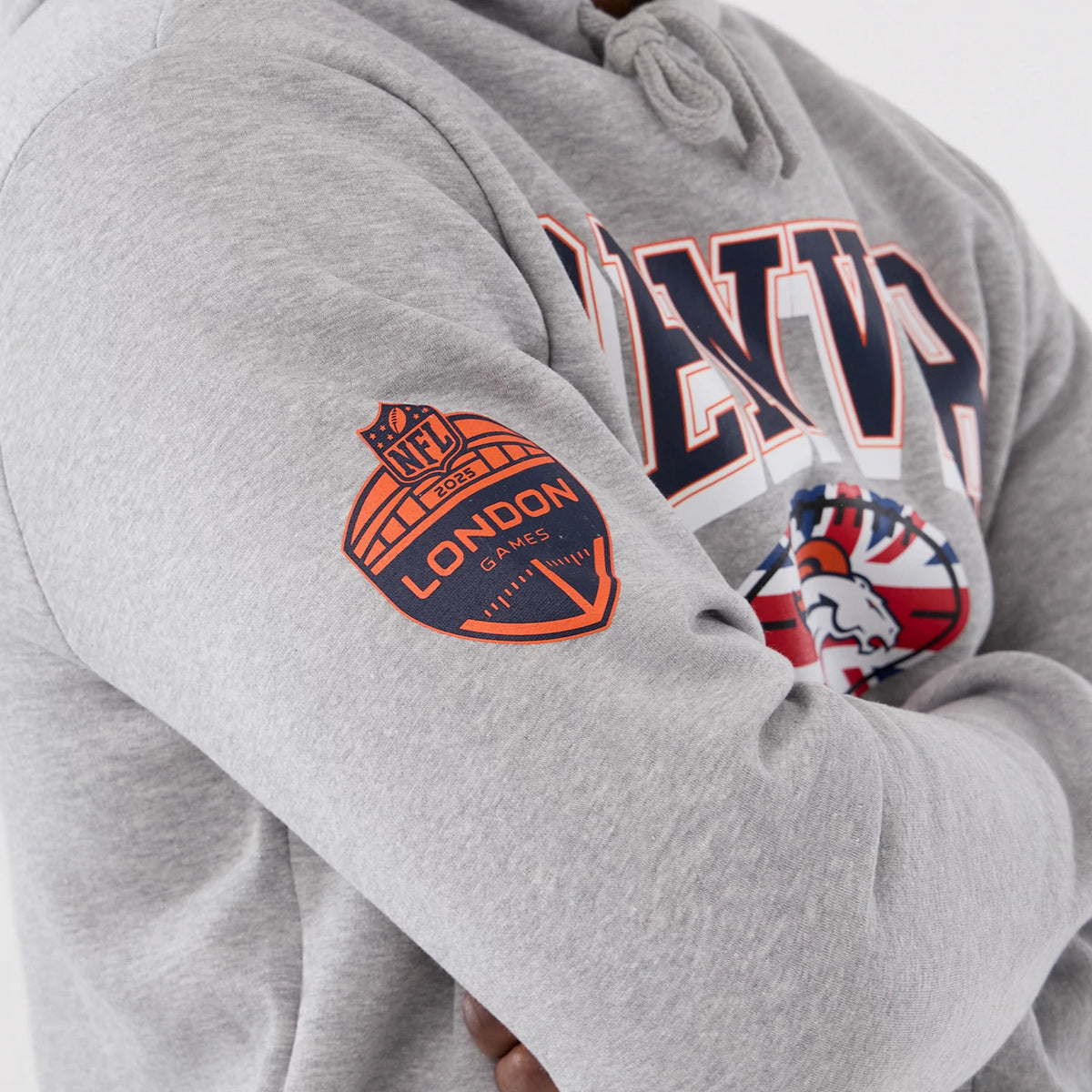 New Era Denver Broncos NFL International Series Games 2025 Grey Pullover Hoodie, three-quarter view showing an orange printed NFL London Games 2025 badge on the sleeve and navy DENVER lettering across the chest on a grey background.