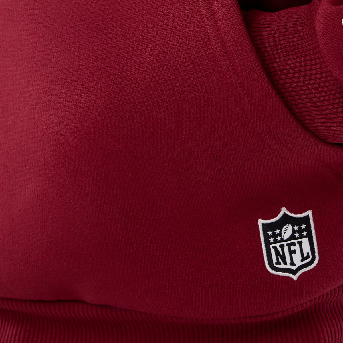 New Era Washington Commanders NFL International Series Games 2025 Dark Red Premium Pullover Hoodie, close-up view showing the ribbed hem and a white embroidered NFL logo near the edge on the dark red fabric.