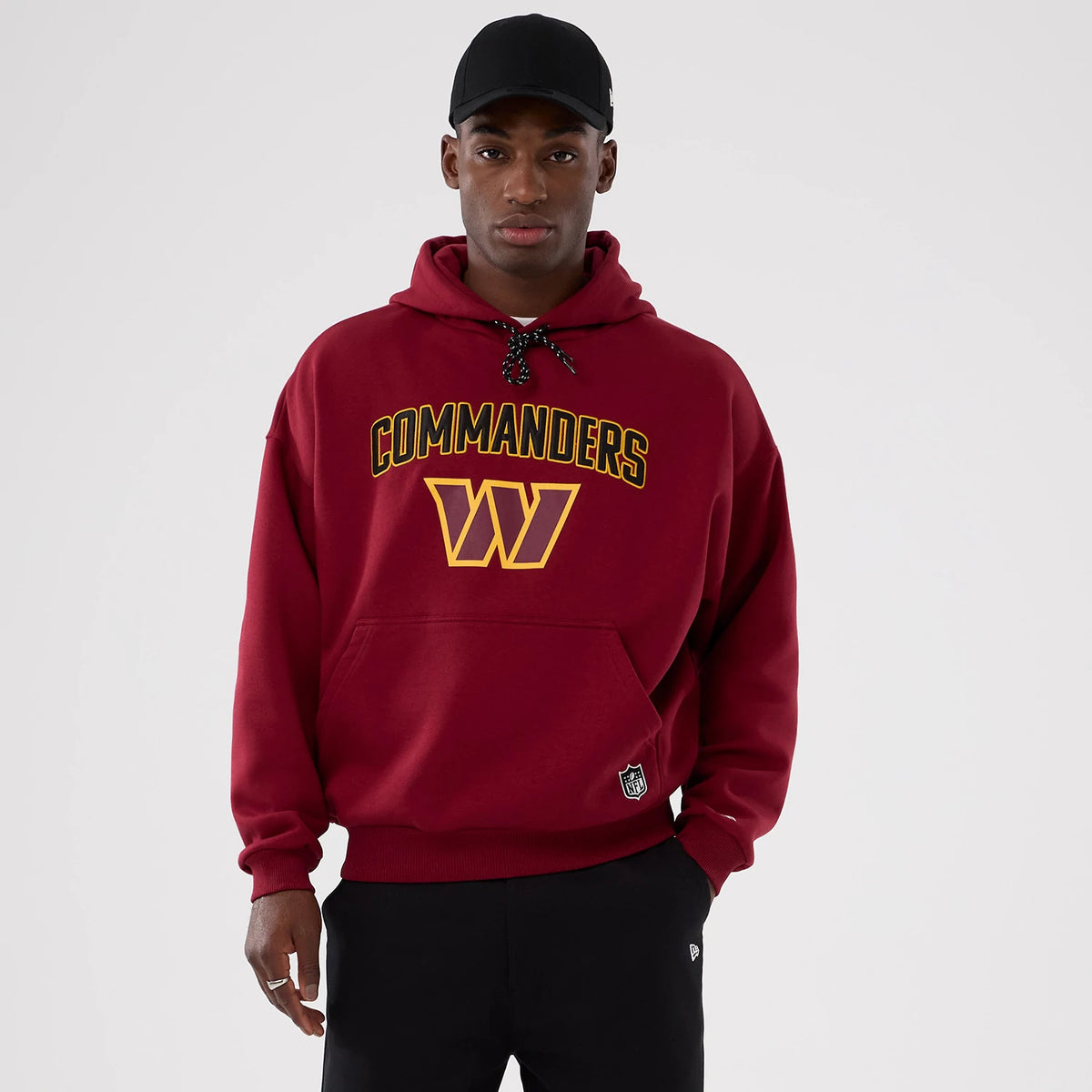 New Era Washington Commanders NFL International Series Games 2025 Dark Red Premium Pullover Hoodie, front view on-model featuring gold Commanders text across the chest with a large matching W logo beneath, embroidered NFL shield logo on the pocket.