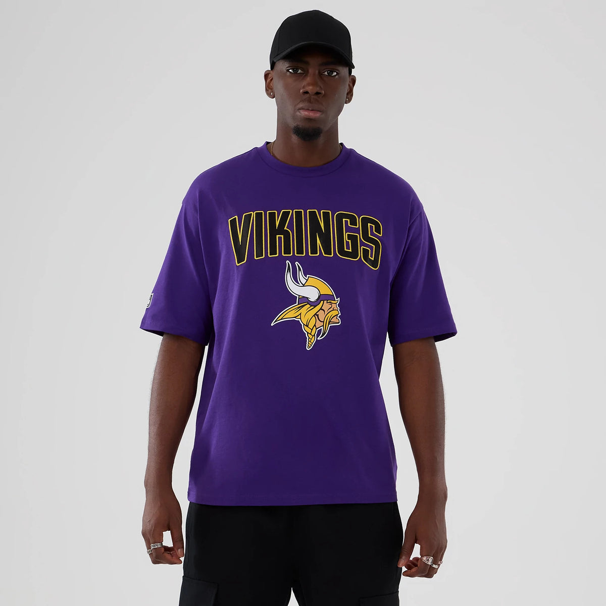 New Era Minnesota Vikings NFL International Series Games 2025 Purple Premium T-Shirt, front view on-model showing yellow VIKINGS lettering and a large embroidered team logo on the chest with a black cap complementing the look.