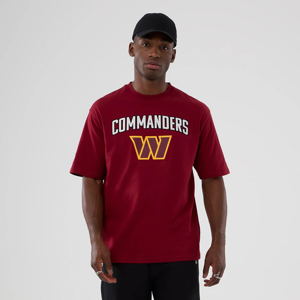 New Era Washington Commanders NFL International Series Games 2025 Dark Red Premium T-Shirt, front view on-model displaying the white printed Commanders lettering and yellow W logo centred on the chest, styled with a plain black cap.