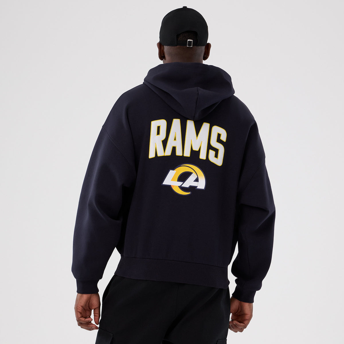 New Era LA Rams NFL International Series Games 2025 Navy Premium Pullover Hoodie, back view on a model wearing a black cap, featuring RAMS lettering and the LA logo in white and gold on a dark navy background.