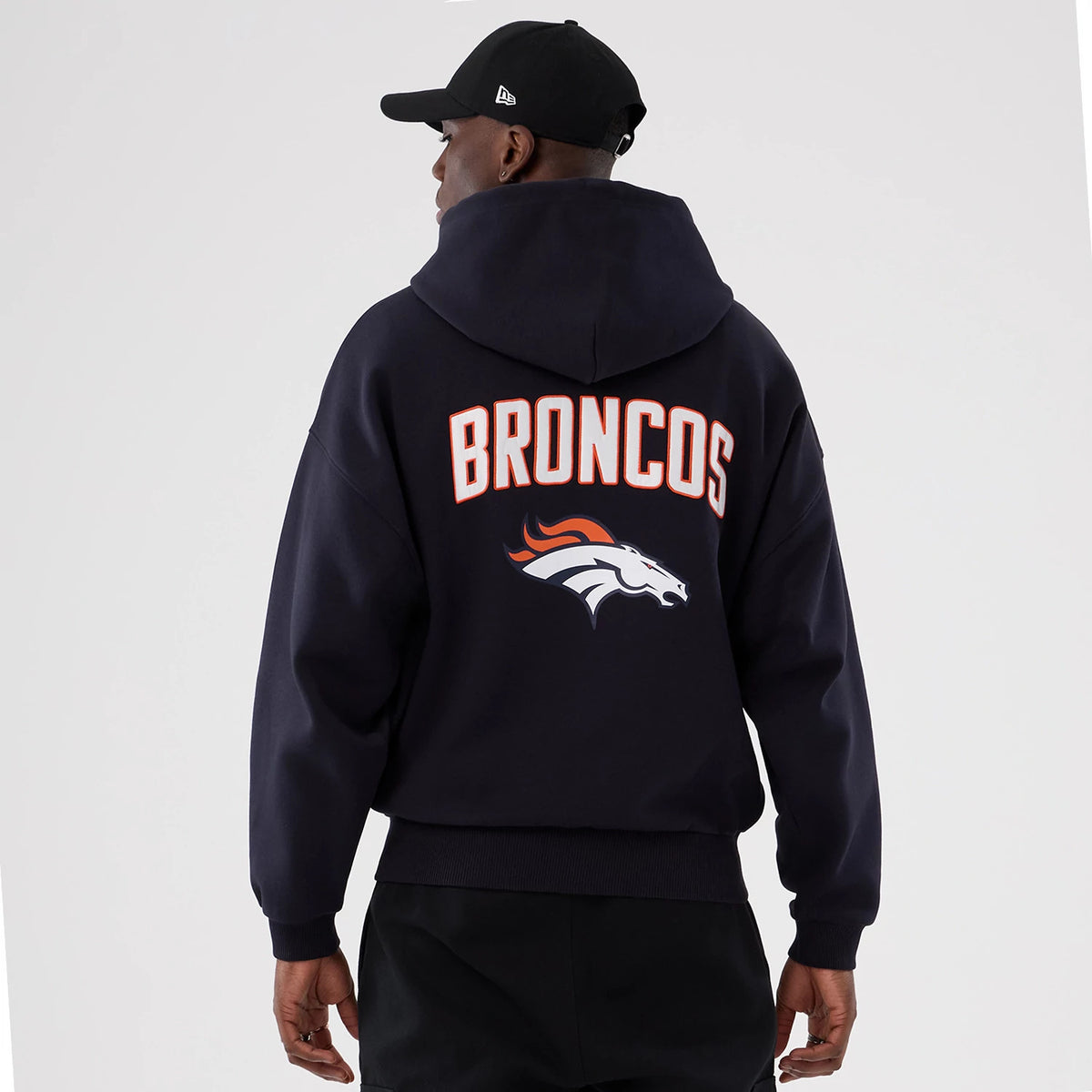 New Era Denver Broncos NFL International Series Games 2025 Navy Premium Pullover Hoodie, back view featuring the white and orange Broncos lettering across the upper back above the team logo, shown on a model styled with a black cap.