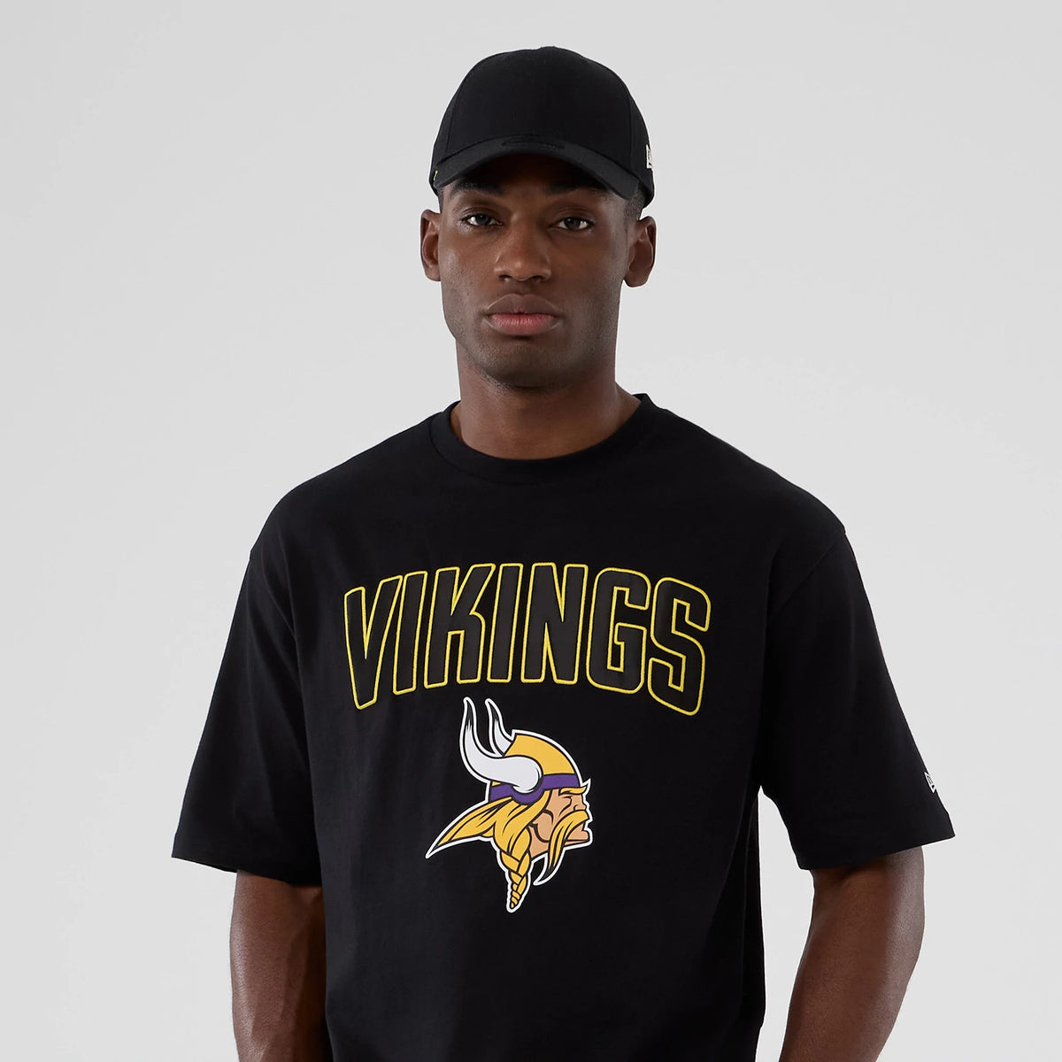 New Era Minnesota Vikings NFL International Series Games 2025 Black Premium T-Shirt, front view on a model showing VIKINGS text in yellow and the team's logo beneath it, worn with a plain black cap.