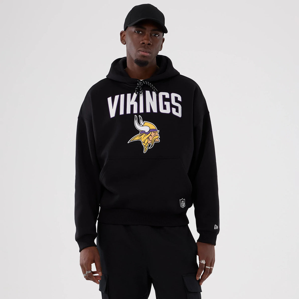 New Era Minnesota Vikings NFL International Series Games 2025 Black Premium Pullover Hoodie, front view on a model showing the Vikings lettering and logo printed across the chest, with an embroidered New Era logo near the cuff.