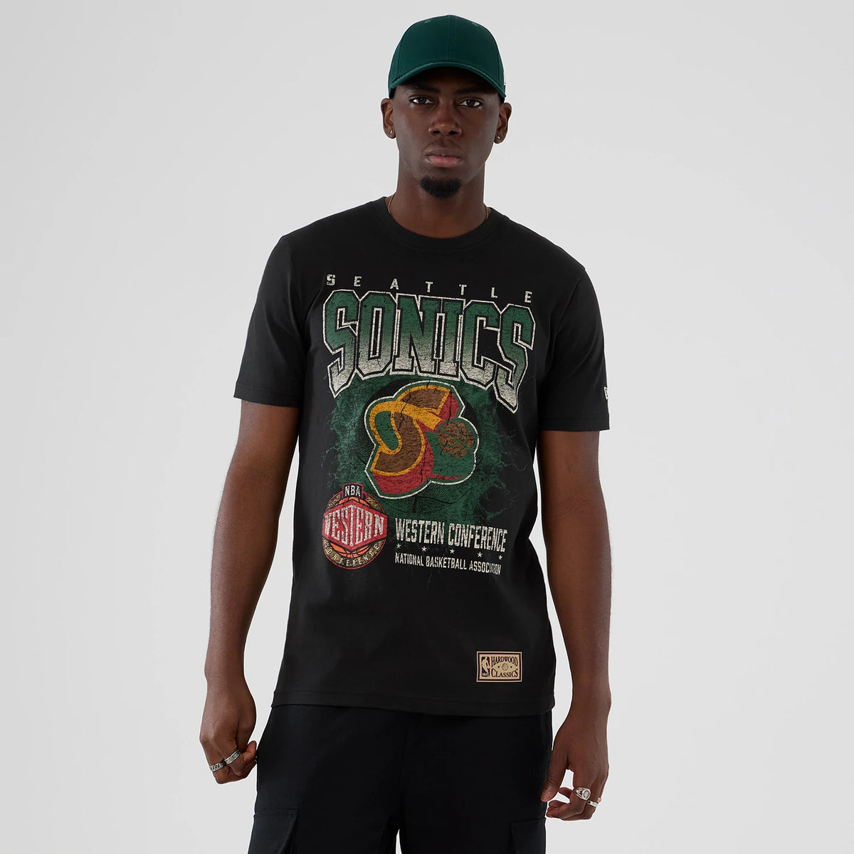 New Era Seattle Supersonics NBA Hardwood Classic Black T-Shirt, front view with large printed Sonics graphic centred on the chest, Western Conference emblem below, worn by a model with a dark green cap.