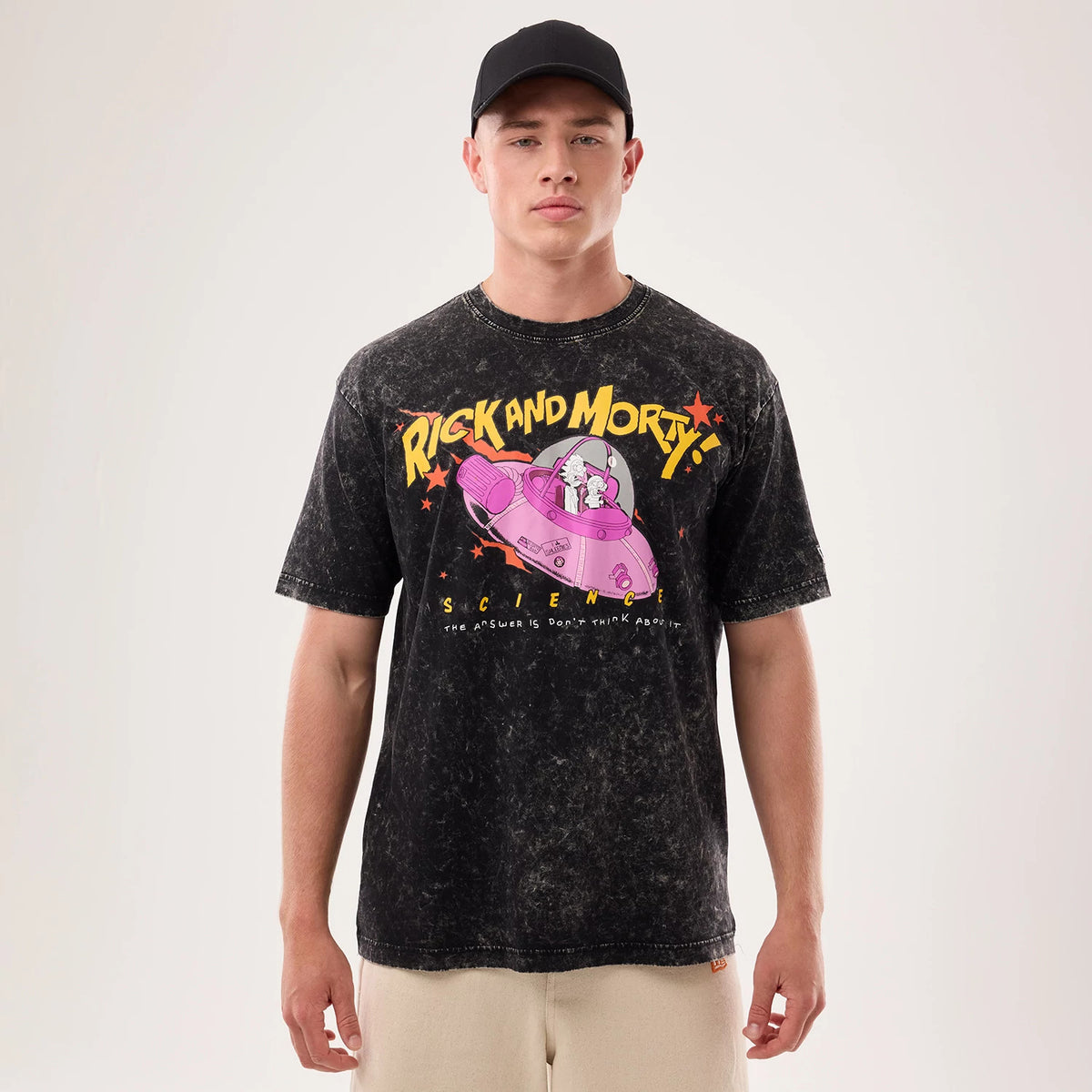 New Era Rick And Morty Science Black T-Shirt, front view on-model featuring a printed graphic of Rick and Morty in a pink spaceship with yellow and red stars and lettering, styled with neutral coloured trousers and a plain black cap.