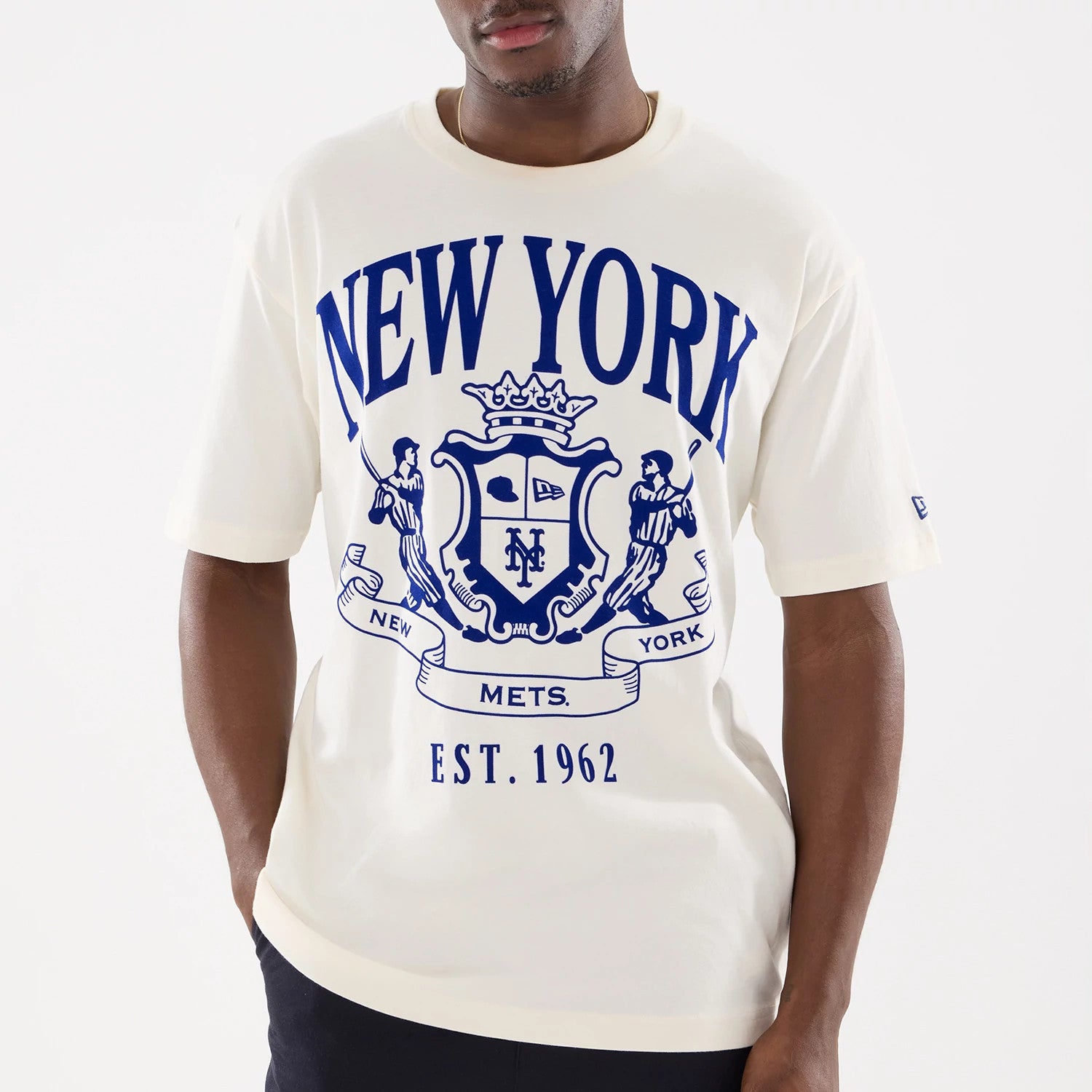 New Era New York Mets MLB Life Graphic Light Beige T-Shirt, front view on a model showing a large blue graphic with a crest, New York in letters above it, and the New Era logo embroidered on the left sleeve.