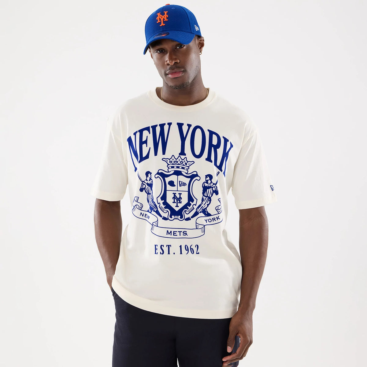 New Era New York Mets MLB Life Graphic Light Beige T-Shirt, front view on a model showing a large blue graphic with NEW YORK lettering and a crest featuring the New Era logo with Est. 1962, paired with a blue cap displaying an embroidered Mets logo.