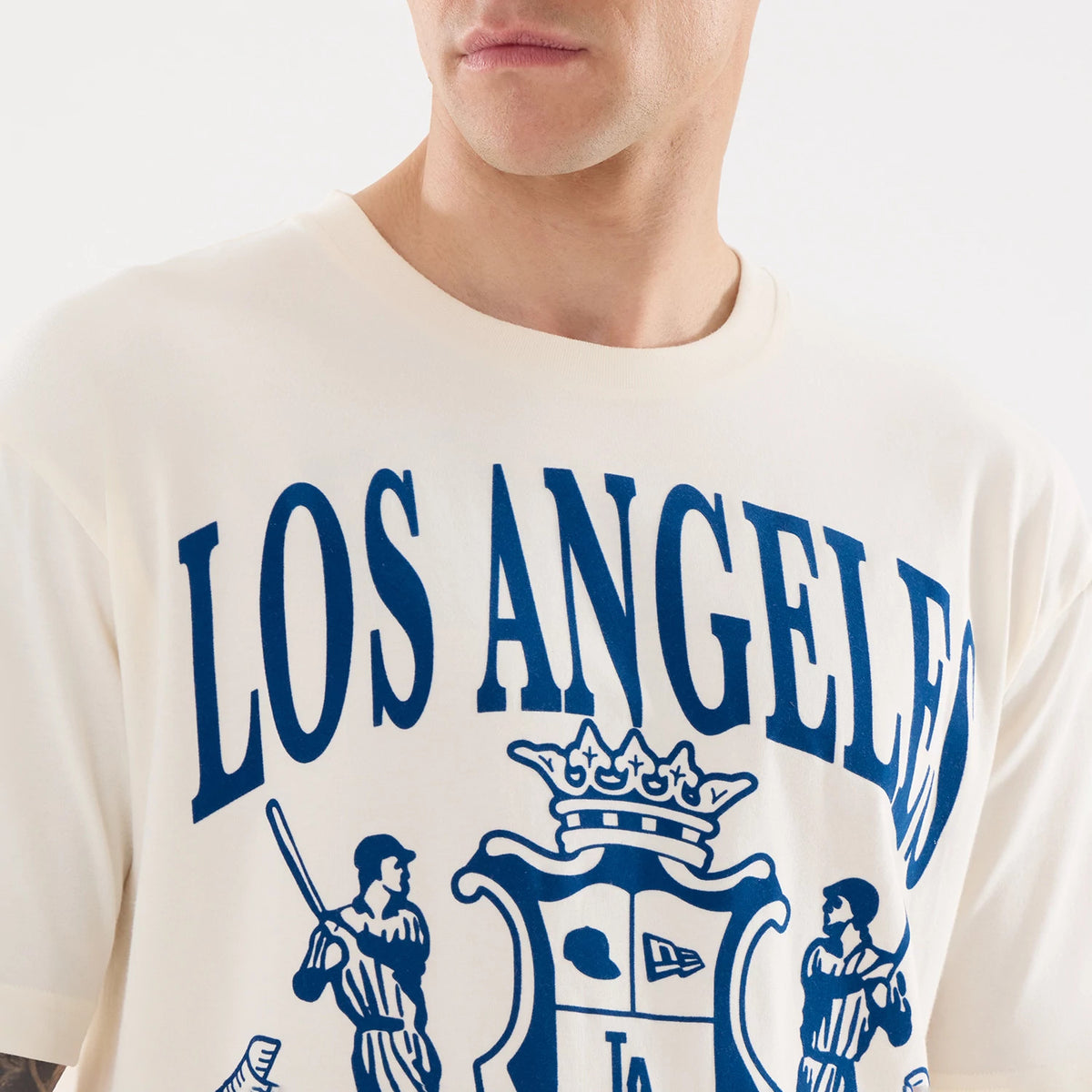 New Era LA Dodgers MLB Life Graphic Light Beige T-Shirt, front view on a model, features large blue Los Angeles lettering across the chest with a central graphic depicting two baseball players and a crest, all in an oversized light beige design.