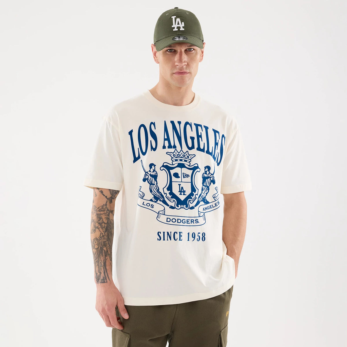 New Era LA Dodgers MLB Life Graphic Light Beige T-Shirt, front view on a model wearing an olive green cap, featuring large blue Los Angeles text with an elaborate crest graphic centred on the chest and Dodgers Since 1958 text along the bottom.