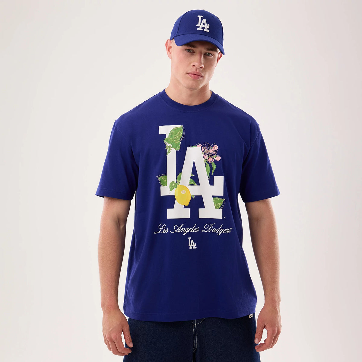 New Era LA Dodgers MLB Life Graphic Dark Blue T-Shirt, front view on a model wearing a matching blue cap, showing a large white LA logo with floral and lemon graphic elements, and Los Angeles Dodgers in script lettering below.