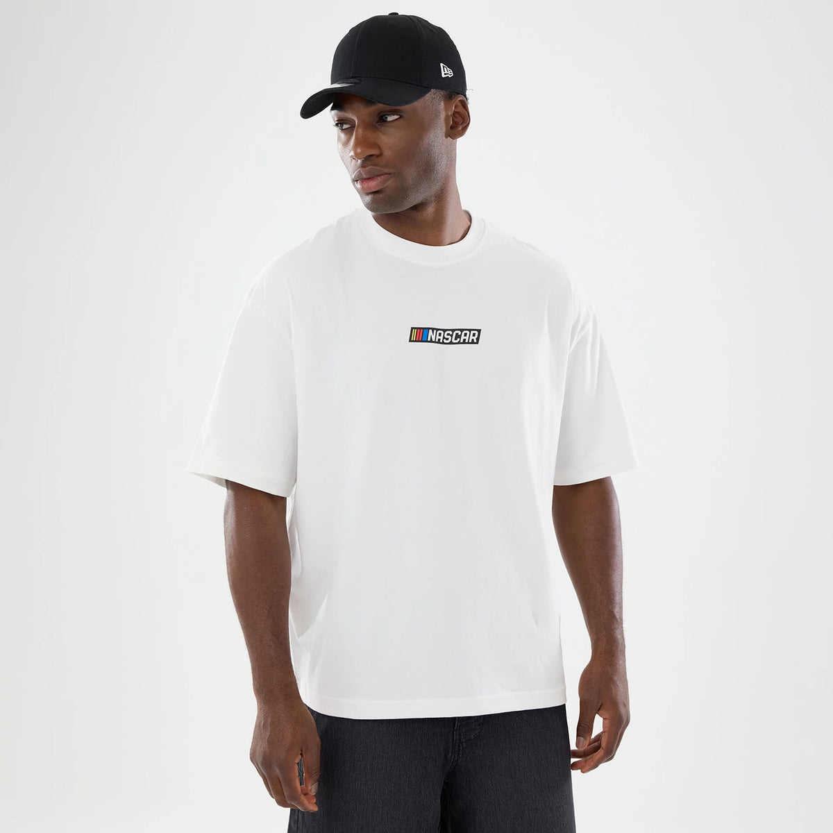 New Era Nascar Logo White T-Shirt, front view on a model wearing a plain black cap, displaying the multicoloured embroidered Nascar logo prominently on the chest of the white t-shirt, short sleeves and a crew neckline adding to the casual style.