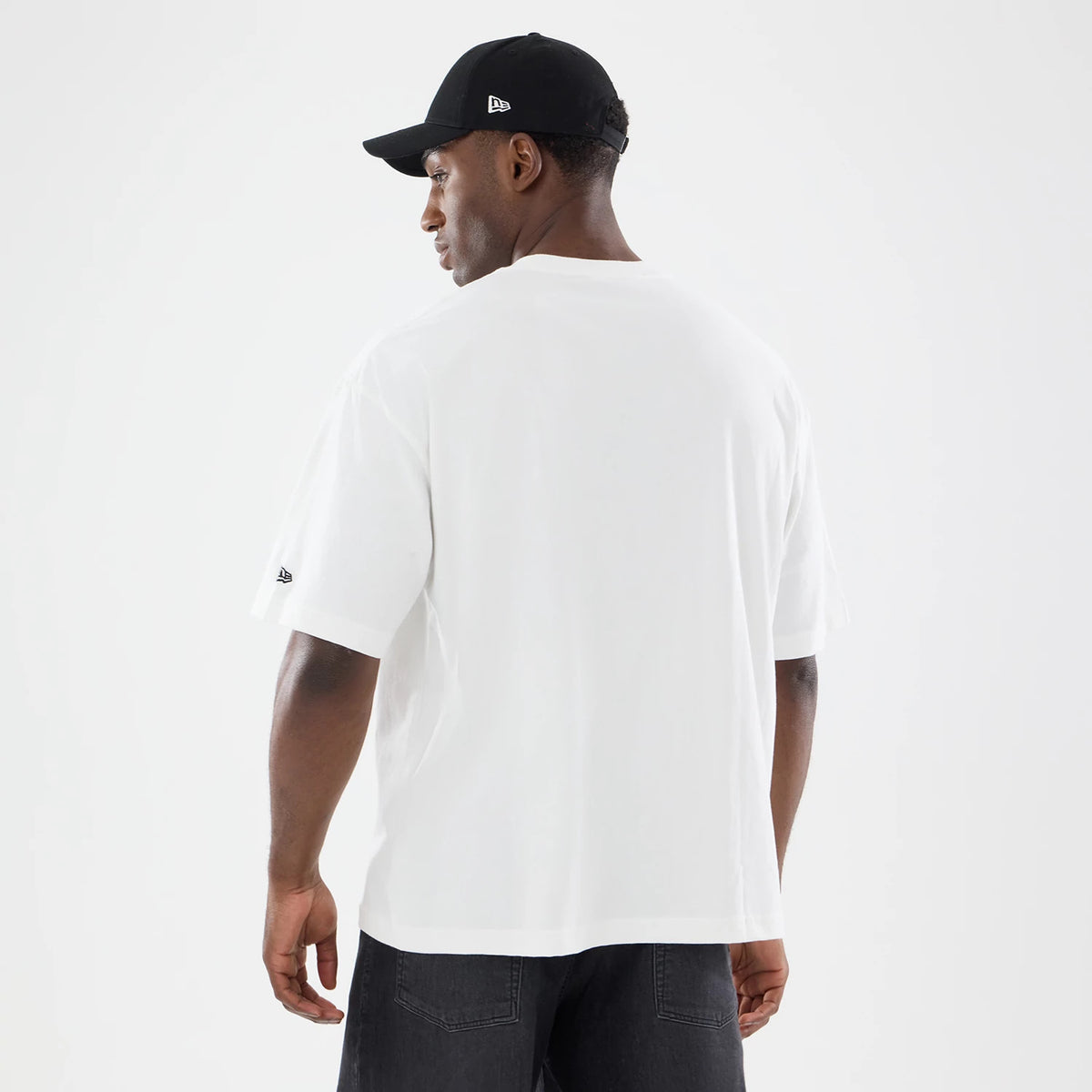 New Era Nascar Logo White T-Shirt, back view on-model with a black cap, showcasing a clean white design, a small New Era embroidered logo on the left sleeve, and a relaxed fit over dark jeans for a casual look.