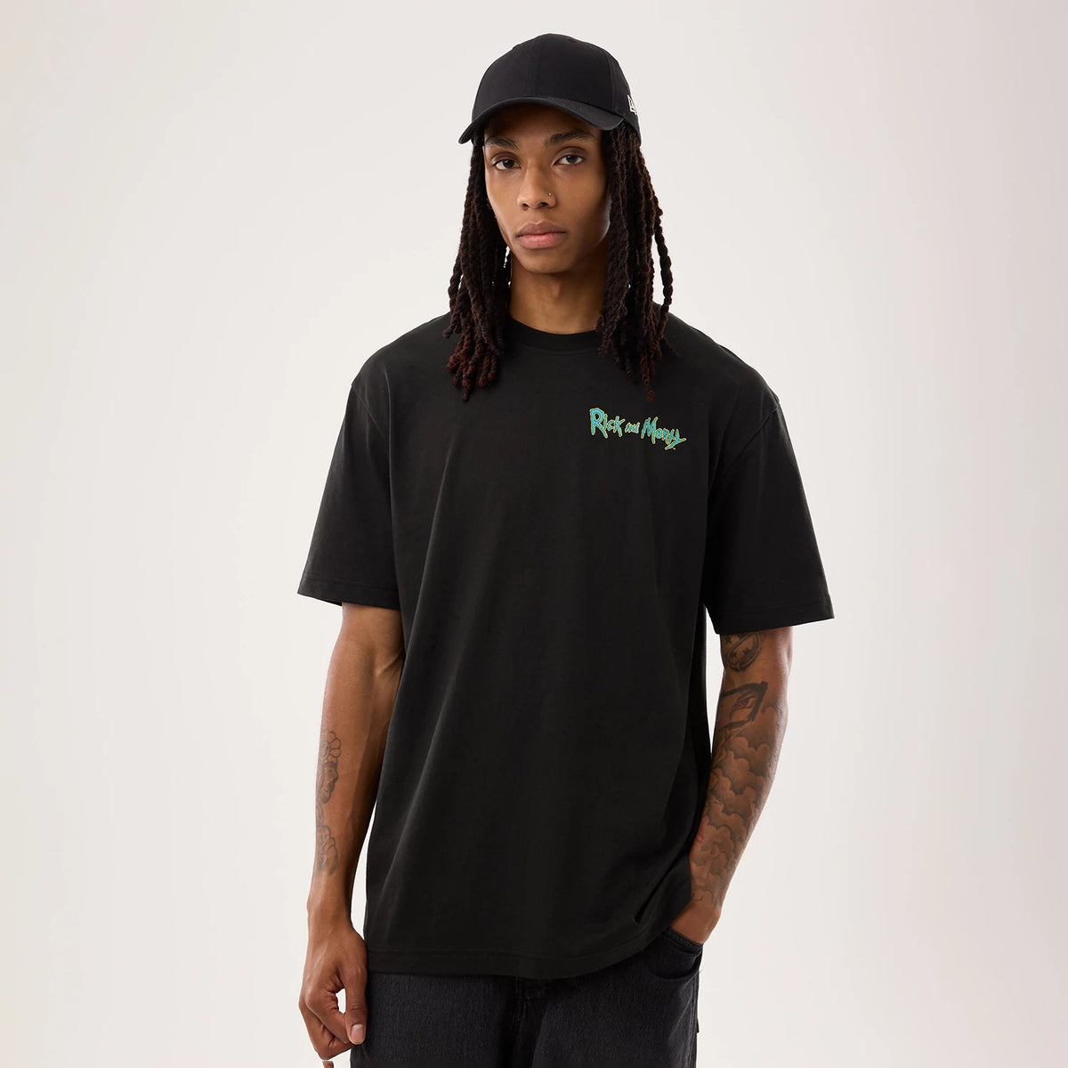 New Era Rick And Morty Portal Gun Black T-Shirt, front view on a model wearing a black cap, featuring a small green embroidered Rick and Morty logo on the left chest, short sleeves and a relaxed fit in solid black fabric.