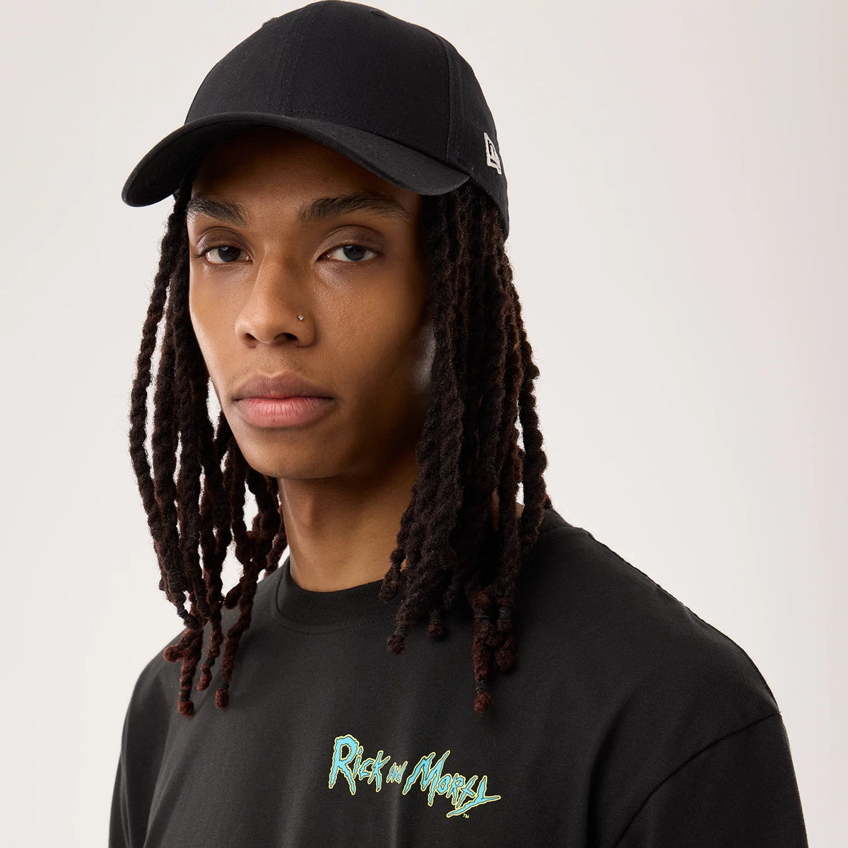 New Era Rick And Morty Portal Gun Black T-Shirt, front view on a model, featuring the Rick and Morty logo printed on the chest in green, styled with a plain black New Era cap displaying an embroidered New Era logo on the side.