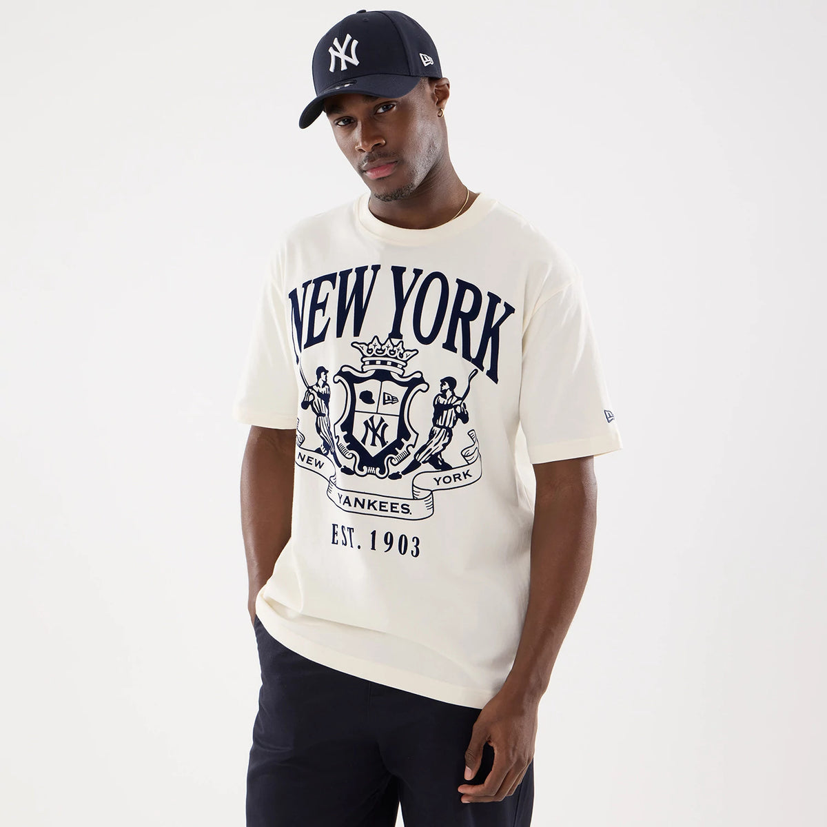 New Era New York Yankees MLB Life Graphic Light Beige T-Shirt, front view on-model showcasing a large dark blue printed New York Yankees logo with crest and EST. 1903 text, with the New Era logo embroidered on the left sleeve.
