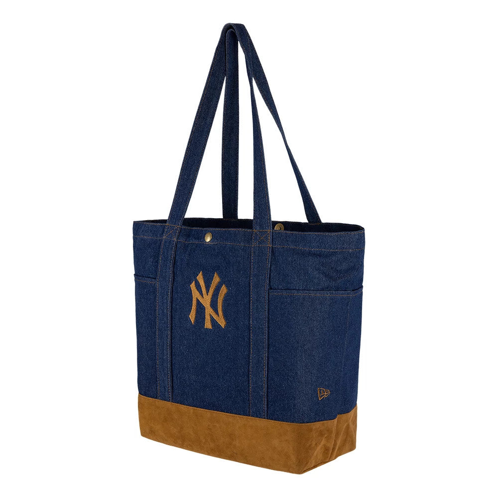 New Era New York Yankees MLB Suede Denim Navy Tote Bag, three-quarter view showing the embroidered Yankees logo in the centre of the denim panel, long navy handles attached at the top, and a brown suede strip along the base of the bag.