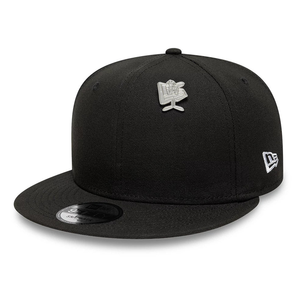 New Era Flag Character Silver Pin Badge, front view on a black cap showing a silver pin badge on the structured crown, an embroidered New Era logo on the side, and a curved black visor with a sticker on the top.